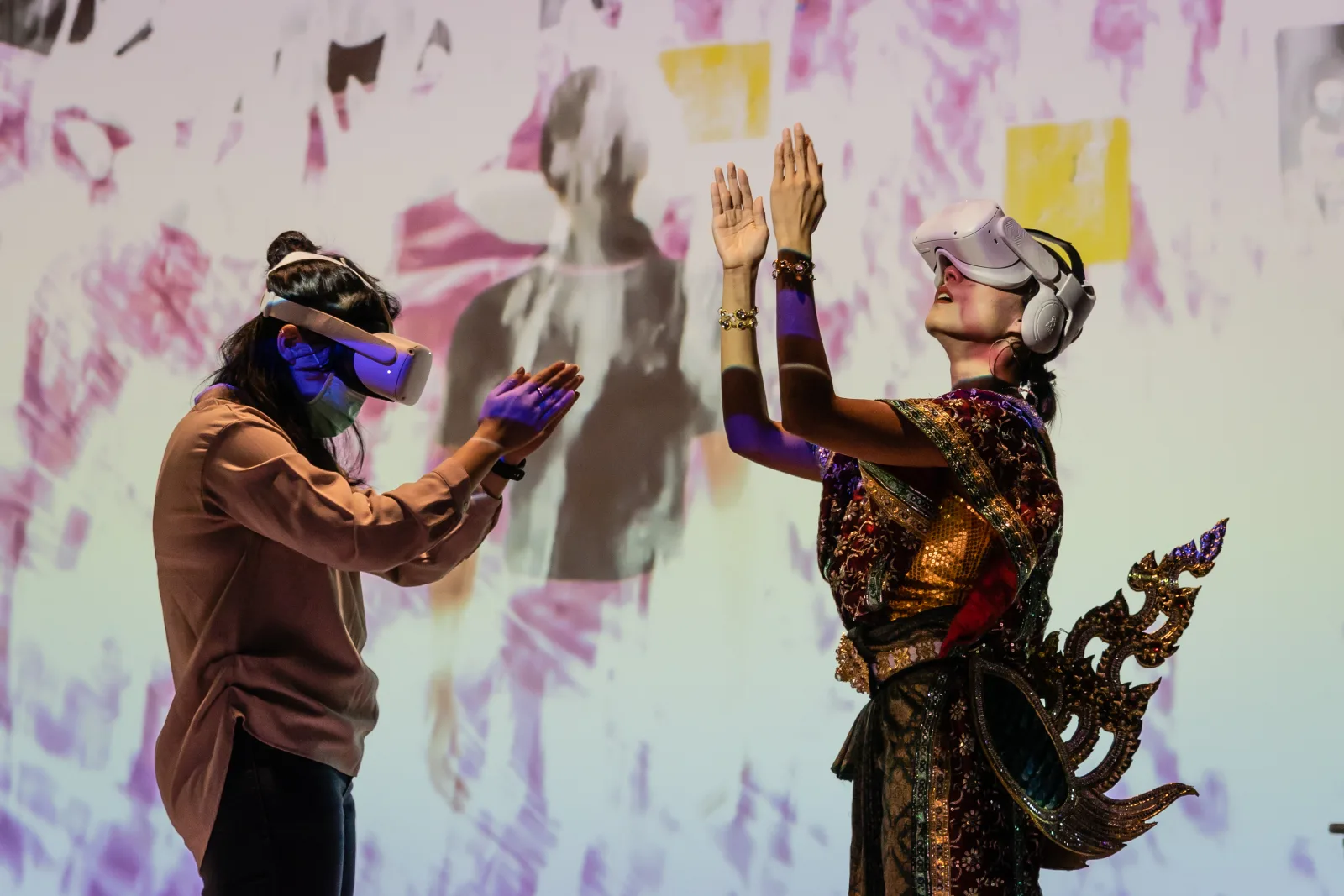 A dancer in Thai costume wearing a VR headset raises her hands in a wai, facing a participant in a headset who mirrors the gesture, against a projected wall of flowers.